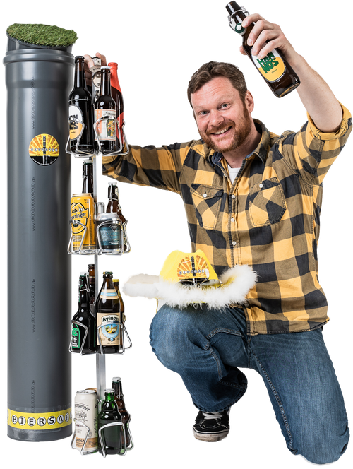 Jens Meyer, of BIERSAFE, grins as he holds the scaffold of a BIERSAFE filled with beer bottles and lifts a beer bottle with his other hand. A party hat is placed on his knee. Next to him is a BIERSAFE tube with an artificial grass lid.